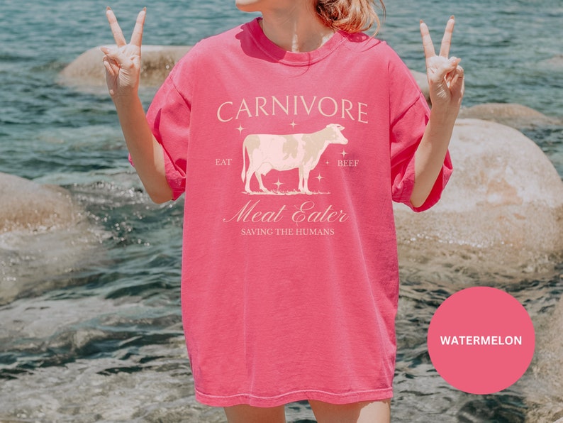 May include: Pink t-shirt with a white cow graphic and the text "CARNIVORE Eat Beef Meat Eater Saving the Humans" on the front. The shirt is being worn by a person standing in front of a body of water.