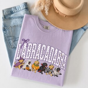 May include: A light purple t-shirt featuring the text "LABRACADABRA" in white with purple polka dots. Below the text are illustrations of six dogs in various Halloween costumes, including a wizard, ghost, pumpkin, vampire, skeleton, and mummy.