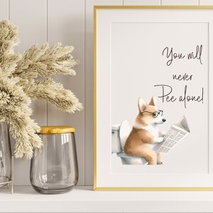 May include: A framed print with a gold border features a watercolor illustration of a corgi wearing glasses and reading a newspaper while sitting on a toilet. The text above the corgi reads, "You will never Pee alone!"