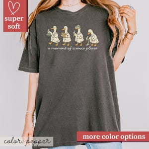 May include: Pepper-colored t-shirt featuring a whimsical design of four ducks in lab coats, each engaged in a different scientific activity. The phrase "a moment of science please" is printed below the ducks. The shirt is labeled "super soft".