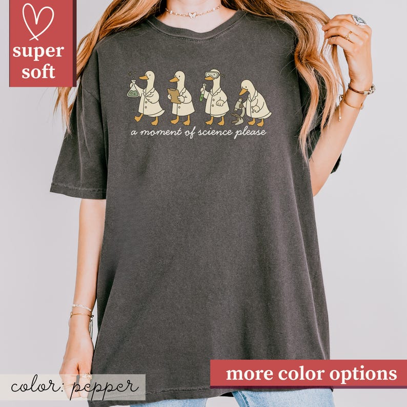 May include: Pepper-colored t-shirt featuring a whimsical design of four ducks in lab coats, each engaged in a different scientific activity. The phrase "a moment of science please" is printed below the ducks. The shirt is labeled "super soft".
