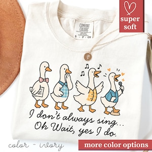 May include: Ivory-colored t-shirt featuring a cartoon design of four geese, each with unique outfits and expressions. The geese are singing, with musical notes and hearts around them. The text "I don't always sing... Oh Wait, yes I do." is printed below.