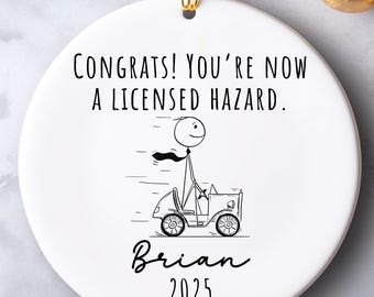 Può includere: Ornamento rotondo in ceramica bianca con un anello dorato per appenderlo. L'ornamento presenta un omino stilizzato alla guida di un'auto, con il testo "CONGRATS! YOU'RE NOW A LICENSED HAZARD." e il nome "Brian" con l'anno "2025".