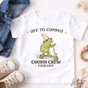 K&ouml;nnte beinhalten: Wei&szlig;es T-Shirt mit einem gr&uuml;nen Frosch auf einem Roller, der einen Zaubererhut tr&auml;gt. Der Text lautet "OFF TO COMMIT" und "COUSIN CREW FOOLERY". Im Hintergrund sind eine Jeansjacke und beige Turnschuhe zu sehen.