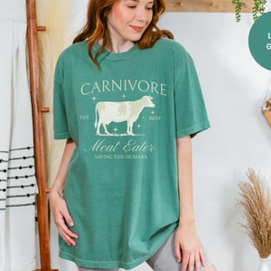 May include: A light green t-shirt with a vintage-style graphic of a cow and the text "CARNIVORE Eat Beef Meat Eater Saving the Humans".