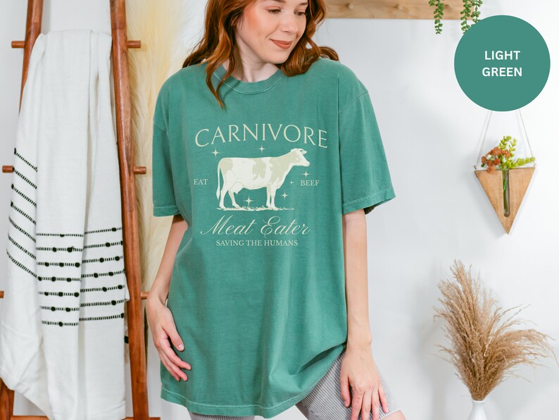 May include: A light green t-shirt with a vintage-style graphic of a cow and the text "CARNIVORE Eat Beef Meat Eater Saving the Humans".
