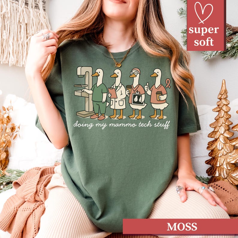May include: Moss green t-shirt featuring a graphic of cartoon ducks in medical attire, with the text "doing my mammo tech stuff." The ducks are depicted with medical equipment and accessories. The shirt has a "super soft" label.