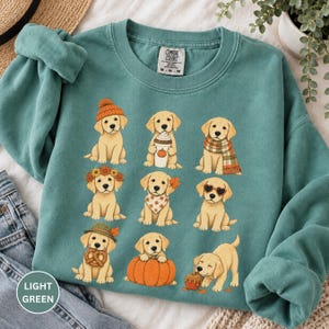 May include: A light green crewneck sweatshirt featuring a graphic print of nine golden retriever puppies in various autumn-themed outfits and poses, including wearing hats, scarves, and holding pumpkins, a pretzel, and a coffee cup with a pumpkin design.