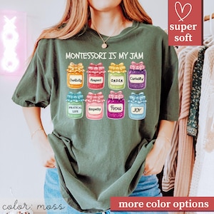May include: Moss green t-shirt with the text "MONTESSORI IS MY JAM" above illustrations of jars labeled with words like "Creativity" and "Joy." The shirt has a relaxed fit and is made of a soft material. The words "super soft" are in a pink box.