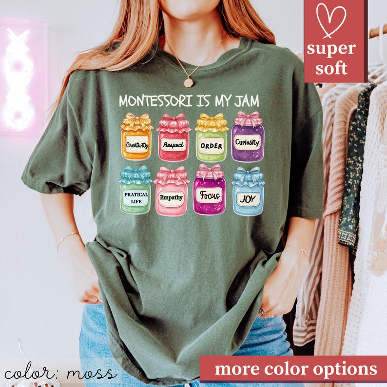 May include: Moss green t-shirt with the text "MONTESSORI IS MY JAM" above illustrations of jars labeled with words like "Creativity" and "Joy." The shirt has a relaxed fit and is made of a soft material. The words "super soft" are in a pink box.