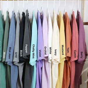 May include: A display of various colored t-shirts hanging on a clothing rack. Colors include light green, moss, blue jean, gray, black, pepper, chalky mint, violet, white, ivory, mustard, yam, cumin, and berry. Each shirt has a label with its color name.