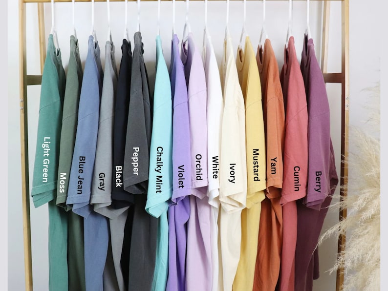 May include: A display of various colored t-shirts hanging on a clothing rack. Colors include light green, moss, blue jean, gray, black, pepper, chalky mint, violet, white, ivory, mustard, yam, cumin, and berry. Each shirt has a label with its color name.