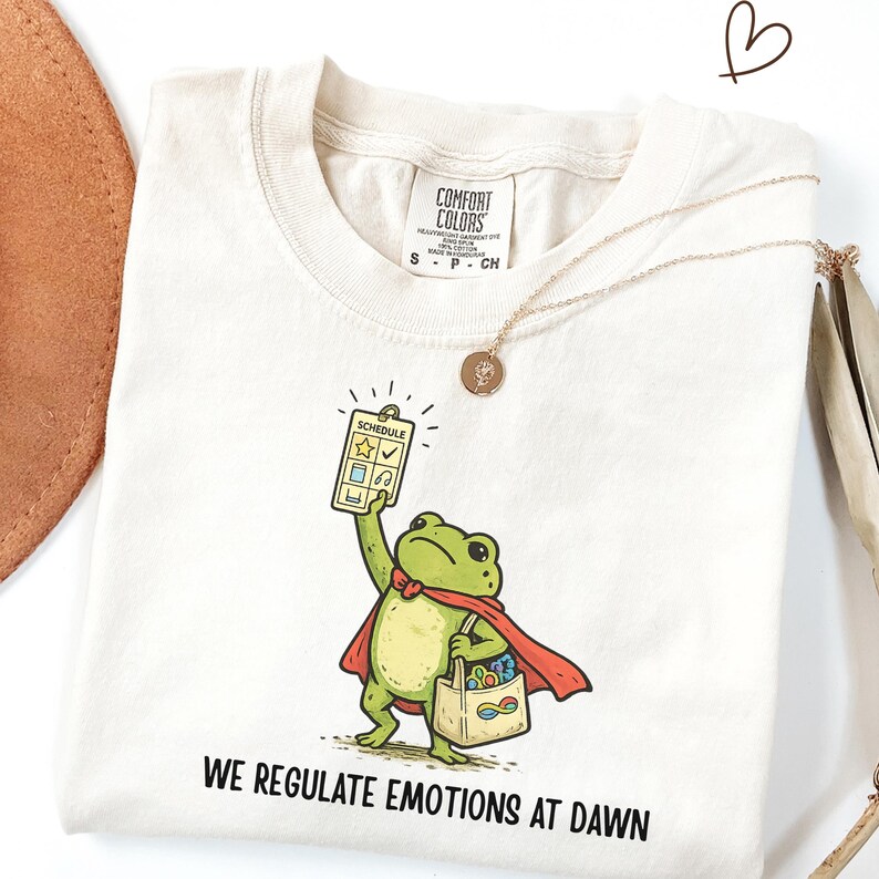 May include: Cream-colored t-shirt with a cartoon frog superhero holding a schedule and a bag. The frog wears a red cape and a bow tie. The text "WE REGULATE EMOTIONS AT DAWN" is printed at the bottom.