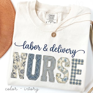 May include: Ivory-colored t-shirt with the words "labor & delivery NURSE" in navy blue script and patchwork letters. The letters have floral and gingham patterns. A gold necklace with a pendant is draped on the shirt.