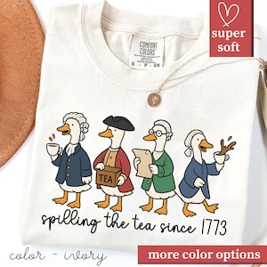 Op de afbeelding: Ivoor kleurig T-shirt met een grappig ontwerp van vier cartoon eenden in koloniale kleding, elk met thee-gerelateerde attributen. De tekst "spilling the tea since 1773" is eronder gedrukt. Het T-shirt heeft een ronde hals en korte mouwen.