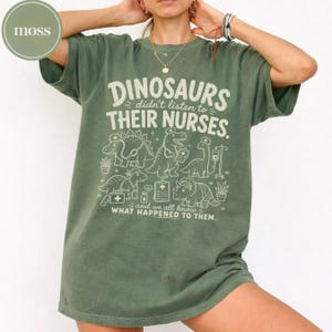 May include: A moss green short-sleeve t-shirt featuring a cream-colored graphic of dinosaurs dressed as medical staff. The text reads: DINOSAURS didn't listen to THEIR NURSES. and we all know WHAT HAPPENED TO THEM. The design includes medical icons.