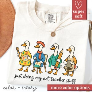 May include: Ivory t-shirt with a cartoon design of four ducks in art teacher attire. The ducks are depicted with a paintbrush, palette, easel, pencil, and pottery. The text "just doing my art teacher stuff" is printed below the ducks.