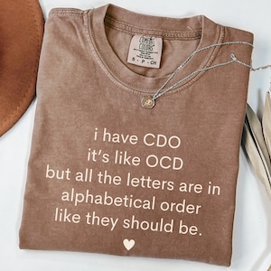 May include: A brown t-shirt with a white graphic that reads "I have CDO it's like OCD but all the letters are in alphabetical order like they should be."  The shirt has a small white heart at the bottom.