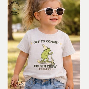 K&ouml;nnte beinhalten: Ein hellgraues T-Shirt mit dem Text "OFF TO COMMIT" und "COUSIN CREW FOOLERY". Das Shirt zeigt einen Cartoon-Frosch, der Roller f&auml;hrt. Der Frosch tr&auml;gt einen Hut und eine Sonnenbrille. Das Shirt wird von einem Kind getragen.