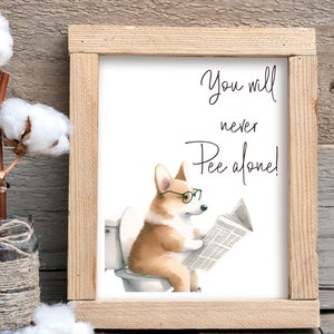 May include: A framed print featuring a watercolor illustration of a corgi wearing glasses and reading a newspaper while sitting on a toilet. The text reads "You will never Pee alone!" The frame is made of light-colored wood.