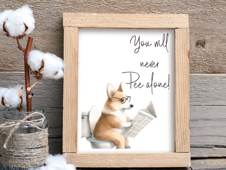 May include: A framed print featuring a watercolor illustration of a corgi wearing glasses and reading a newspaper while sitting on a toilet. The text reads "You will never Pee alone!" The frame is made of light-colored wood.