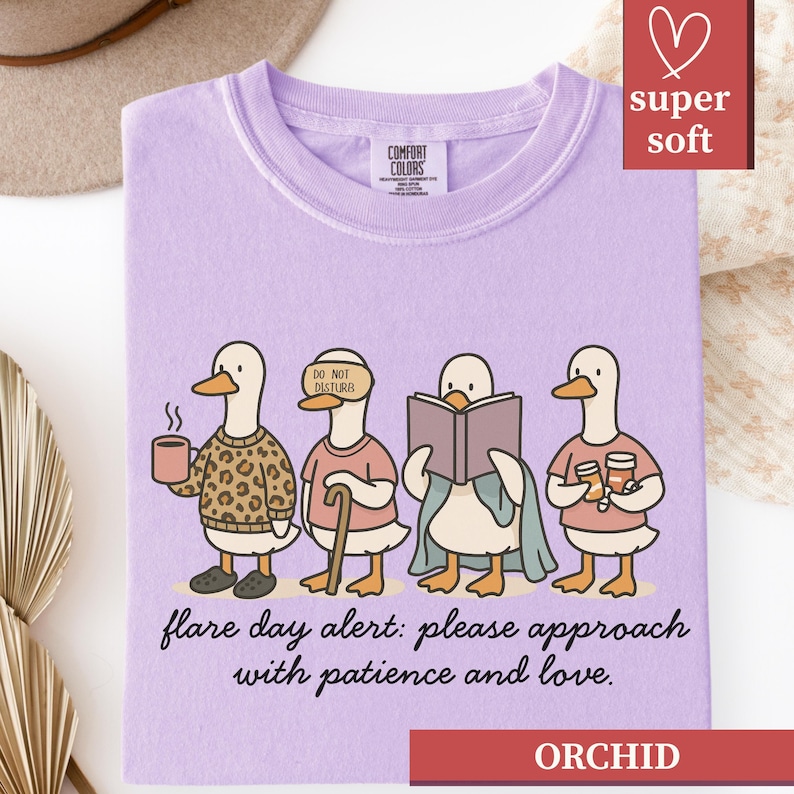 May include: Orchid-colored t-shirt with a graphic of four ducks, each with a unique accessory, and the text "flare day alert: please approach with patience and love." The shirt is labeled "super soft."