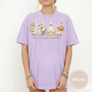 May include: A lilac t-shirt featuring a cartoon design of four ducks involved in baking. The ducks are depicted with jars, aprons, and bread. The phrase "don't even get me started" is printed beneath the ducks in a handwritten style font.