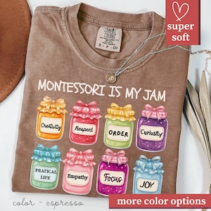 May include: Espresso-colored t-shirt with the text "MONTESSORI IS MY JAM" and colorful jam jar graphics. Each jar has a different label, including "Creativity," "Respect," and "Order." The shirt has a "super soft" label.
