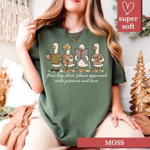 May include: Moss green t-shirt featuring a whimsical illustration of four ducks, each with a unique accessory, holding a coffee cup, cane, book, and drinks. The text "flare day alert: please approach with patience and love" is printed below the ducks.