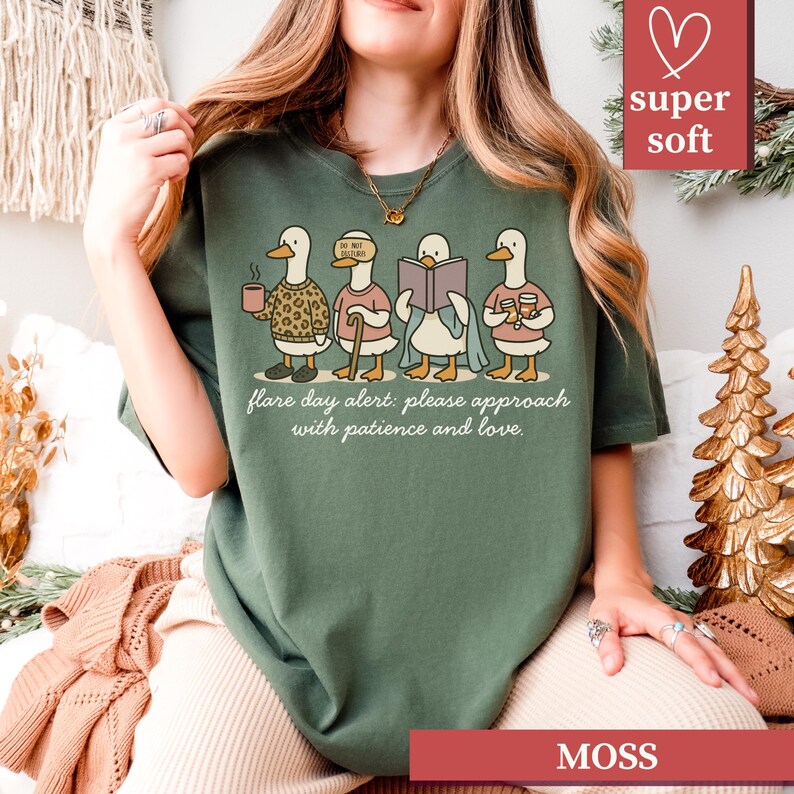 May include: Moss green t-shirt featuring a whimsical illustration of four ducks, each with a unique accessory, holding a coffee cup, cane, book, and drinks. The text "flare day alert: please approach with patience and love" is printed below the ducks.