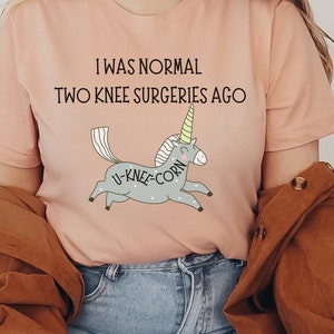 Op de afbeelding: Een roze T-shirt met de tekst "Ik was normaal twee knieoperaties geleden" en een cartoon-einhorn met de tekst "U-knee-corn" aan de zijkant.