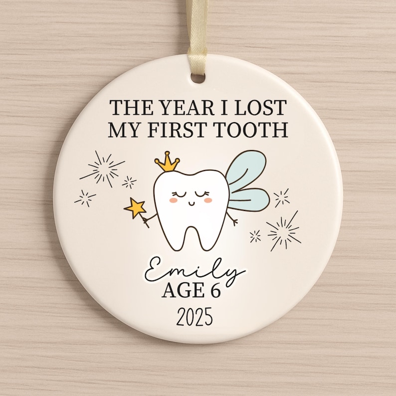 May include: A round, cream-colored ornament with the text "THE YEAR I LOST MY FIRST TOOTH." It features a cartoon tooth with a crown, wings, and a wand. The name "Emily," age 6, and the year 2025 are also printed on the ornament.