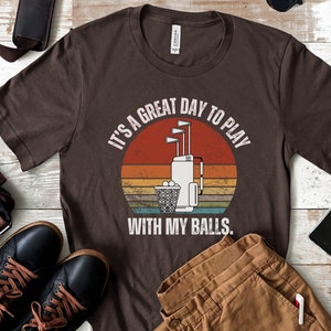 May include: A brown t-shirt with a vintage-style graphic of a golf bag with clubs and golf balls. The text on the shirt reads "It's a great day to play with my balls."