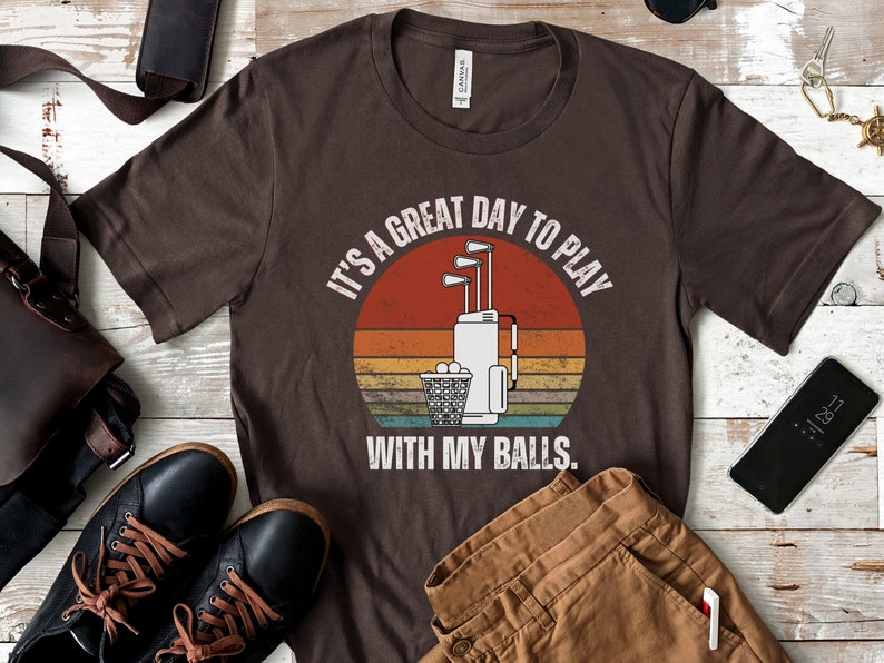 May include: A brown t-shirt with a vintage-style graphic of a golf bag with clubs and golf balls. The text on the shirt reads "It's a great day to play with my balls."