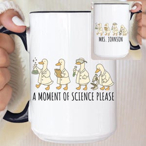 Op de afbeelding: Witte keramische mok met een zwarte rand en handvat. De mok heeft een cartoon illustratie van eenden in laboratoriumjassen, met de tekst "A MOMENT OF SCIENCE PLEASE." Een kleinere mok toont eenden en de naam "MRS. JOHNSON."