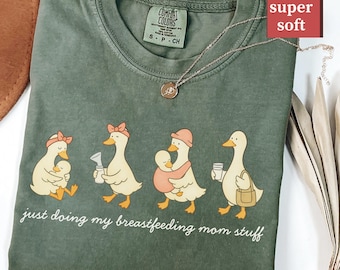 May include: Moss green t-shirt with a whimsical illustration of four ducks, each holding a baby or a bottle. The text "just doing my breastfeeding mom stuff" is printed below the ducks. A gold necklace is draped across the shirt.
