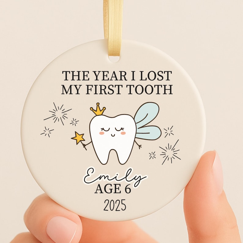 May include: A round, cream-colored ornament with the text "THE YEAR I LOST MY FIRST TOOTH." A cartoon tooth with a crown and wings is featured. The name "Emily," age 6, and the year 2025 are also printed on the ornament. A gold ribbon is attached.