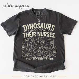 May include: A pepper-colored t-shirt featuring a graphic print with the text "DINOSAURS didn't listen to THEIR NURSES. and we all know WHAT HAPPENED TO THEM." The design includes illustrations of dinosaurs wearing nurse caps and medical gear.