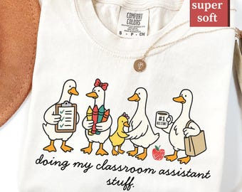 Könnte beinhalten: Elfenbeinfarbenes T-Shirt mit einer Illustration von Enten. Die Enten sind als Klassenassistenten dargestellt, die Gegenstände wie einen Klemmbrett, Buntstifte und eine Kaffeetasse halten. Der Text lautet "doing my classroom assistant stuff."