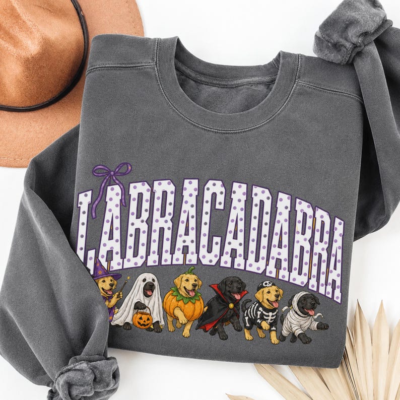 May include: A charcoal grey crewneck sweatshirt featuring a graphic of six Labrador retrievers in various Halloween costumes. The text "LABRACADABRA" is printed in large white letters with purple polka dots above the dogs, accented by a purple bow.