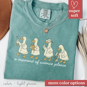 May include: Light green t-shirt with a graphic of four ducks in lab coats, each with science equipment. The text "a moment of science please" is below the ducks. The shirt has a crew neck and short sleeves.