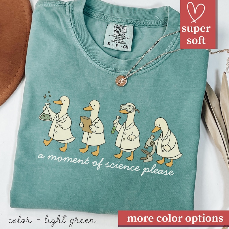 May include: Light green t-shirt with a graphic of four ducks in lab coats, each with science equipment. The text "a moment of science please" is below the ducks. The shirt has a crew neck and short sleeves.