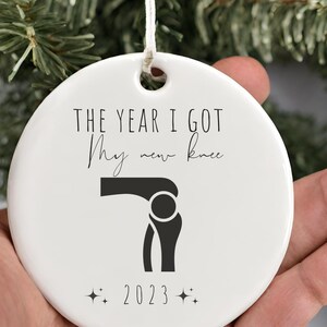 Op de afbeelding: Witte keramische kerstversiering met zwarte tekst die "Het jaar dat ik mijn nieuwe knie kreeg" luidt en een zwart silhouet van een knie. Het jaar 2023 staat onder de knie met twee kleine sterren aan weerszijden.