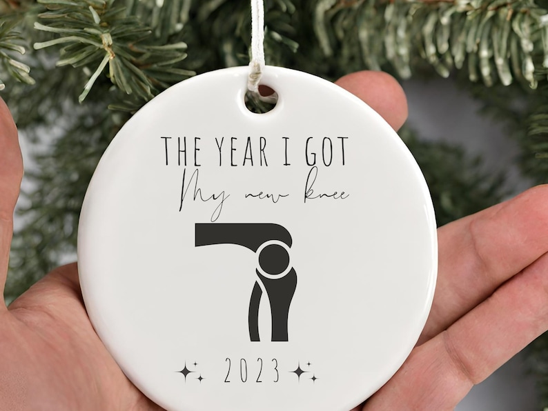 Op de afbeelding: Witte keramische kerstversiering met zwarte tekst die "Het jaar dat ik mijn nieuwe knie kreeg" luidt en een zwart silhouet van een knie. Het jaar 2023 staat onder de knie met twee kleine sterren aan weerszijden.