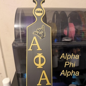 May include: A black and gold paddle with the Greek letters Alpha, Phi, and Alpha. The paddle has a handle with the number 1906 and a hand symbol. The words "Alpha Phi Alpha" are also visible.