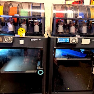 May include: Two black 3D printers with clear tops holding colorful filament spools. Each printer has a display screen and a front door. A yellow emoji sticker is on one printer, and a cartoon sticker is on the other.