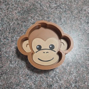 May include: A brown monkey face cookie cutter with a light brown face, black eyes, and a smiling mouth. The cookie cutter is made of plastic and has a detailed design.