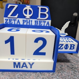 May include: A blue and white desktop calendar with a white dove on top. The calendar is a cube with a drawer that says "ZΦB" and "1920". The top of the calendar says "ZΦB Zeta Phi Beta" and the date is "May 12th".