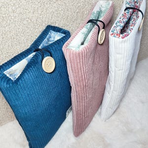 May include: Three corduroy book sleeves in teal, pink, and white. Each sleeve has a contrasting floral lining and a wooden button closure. The sleeves are designed to protect books and are displayed on a fluffy white surface.
