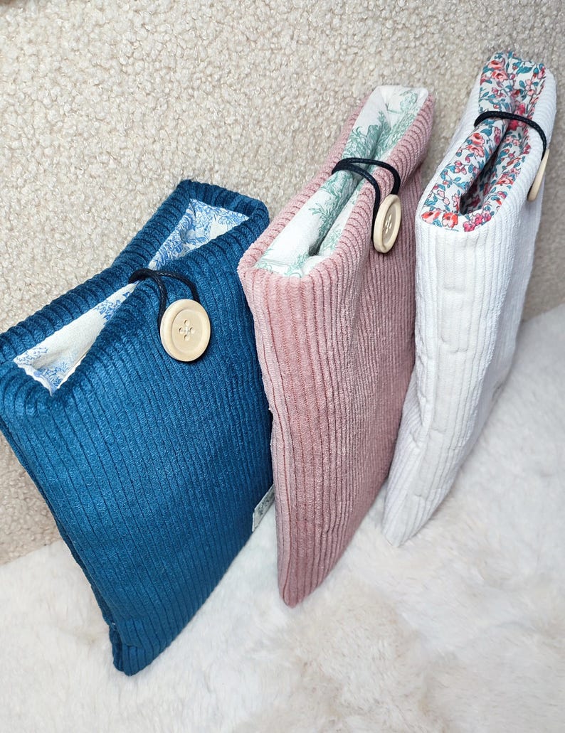 May include: Three corduroy book sleeves in teal, pink, and white. Each sleeve has a contrasting floral lining and a wooden button closure. The sleeves are designed to protect books and are displayed on a fluffy white surface.